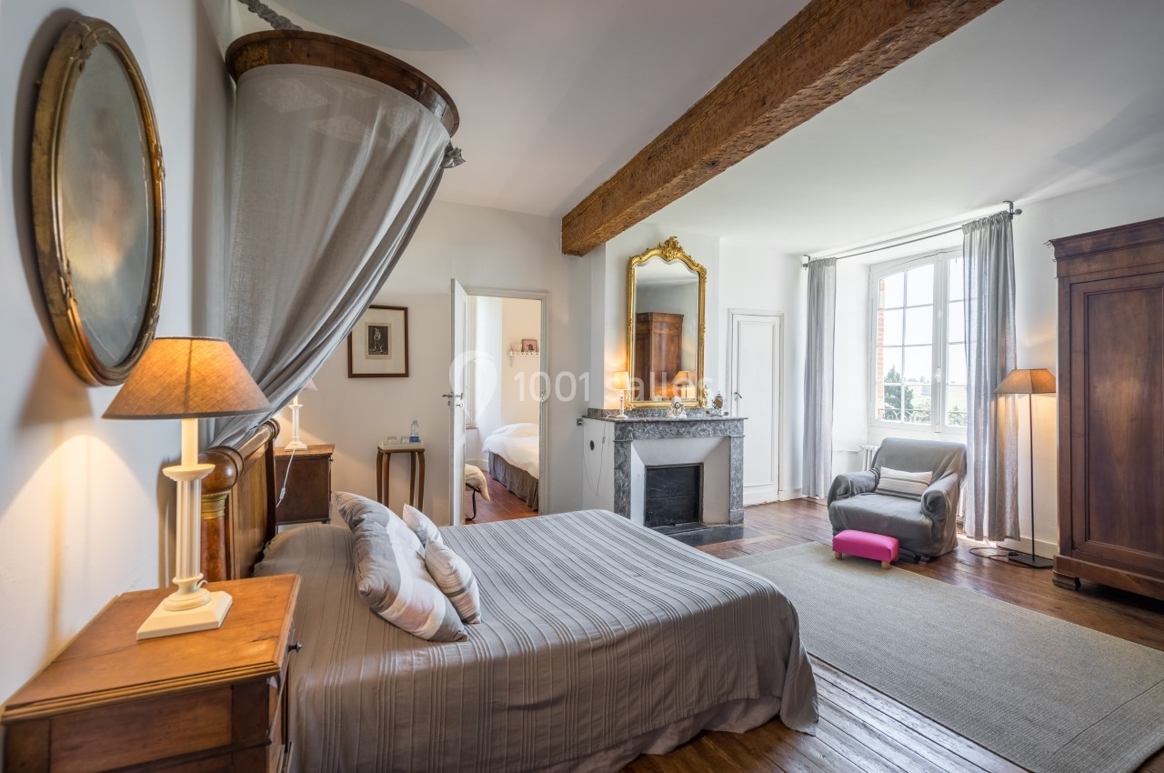 Chambre lumineuse avec lit double, cheminée, armoire en bois, fauteuil et grande fenêtre donnant sur l'extérieur.