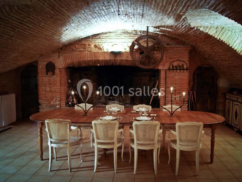 Salle à manger rustique avec table en bois, chaises blanches et cheminée en pierre sous une voûte en briques.