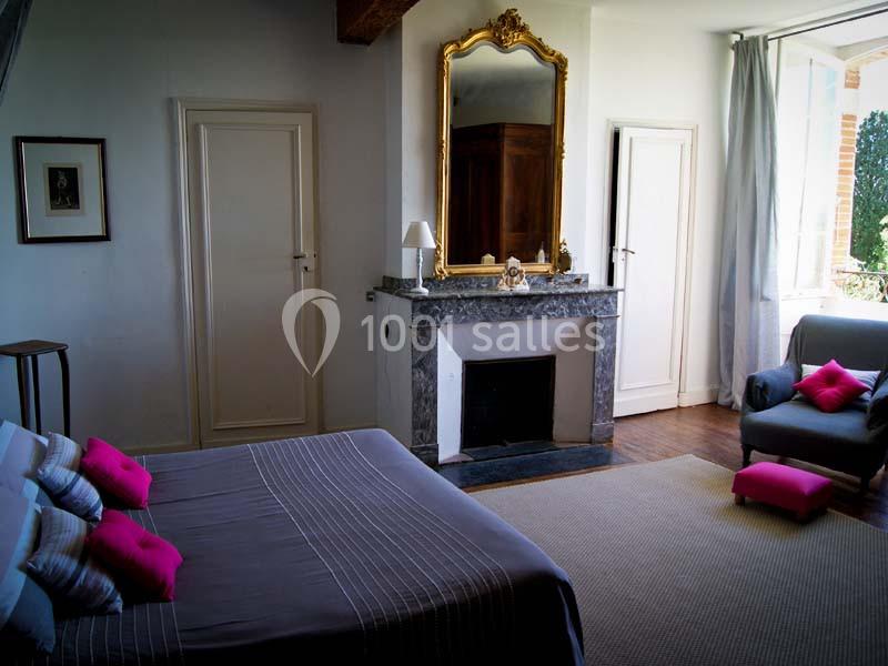 Chambre lumineuse avec lit double, cheminée ornée d'un miroir doré, fauteuil gris et vue sur l'extérieur.
