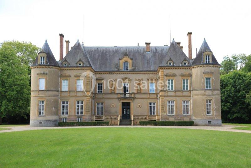 Façade d'un château en pierre beige avec toit en ardoise, entouré de pelouse et d'arbres verdoyants.