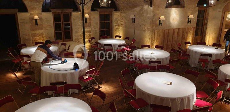 Un homme prépare des tables rondes couvertes de nappes blanches dans une salle en pierre éclairée.