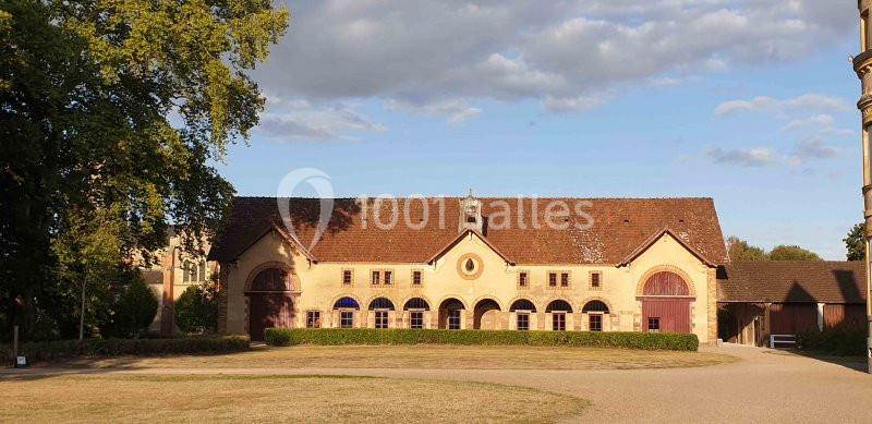 Bâtiment ancien avec toit en tuiles, façade beige et plusieurs arches, entouré d'arbres et d'un espace dégagé.