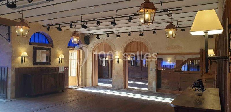 Salle intérieure lumineuse avec murs en pierre, poutres apparentes, lampes suspendues et grandes ouvertures en bois.