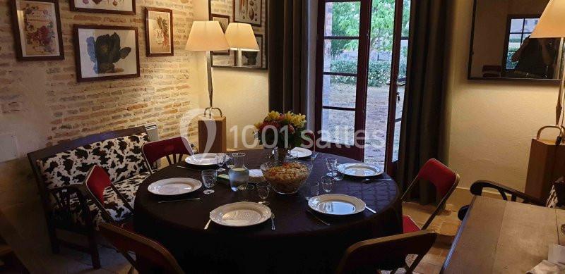 Salle à manger avec table dressée pour quatre personnes, éclairée par des lampes et une porte-fenêtre donnant sur un jardin.
