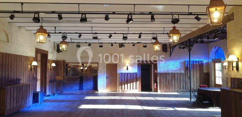 Salle intérieure avec parquet en bois, murs en pierre, éclairage suspendu et lumière naturelle entrant par des fenêtres.