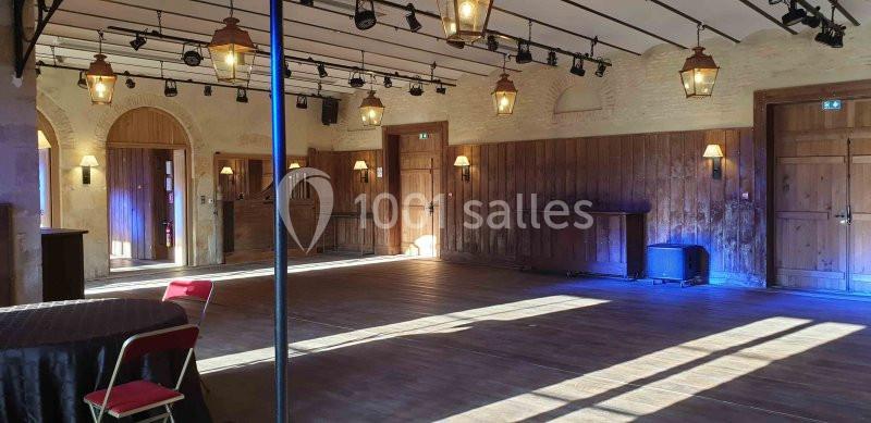 Salle vide avec parquet en bois, murs en pierre, éclairage suspendu et lumière naturelle entrant par les fenêtres.
