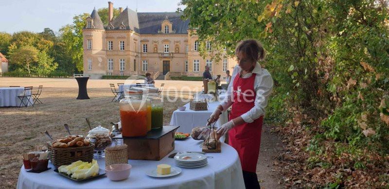 Une personne en tablier rouge découpe de la viande sur une table dressée en extérieur devant un château.