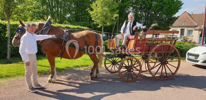 Un homme tient un cheval attelé à une calèche où est assis un autre homme, dans un cadre rural ensoleillé.