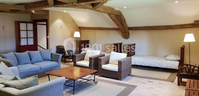 Salon spacieux avec poutres apparentes, canapé, fauteuils en osier, lits simples et éclairage doux.