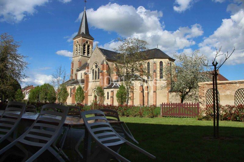 Église en pierre avec clocher, entourée d'arbres et de pelouses, vue depuis des chaises en plein air.