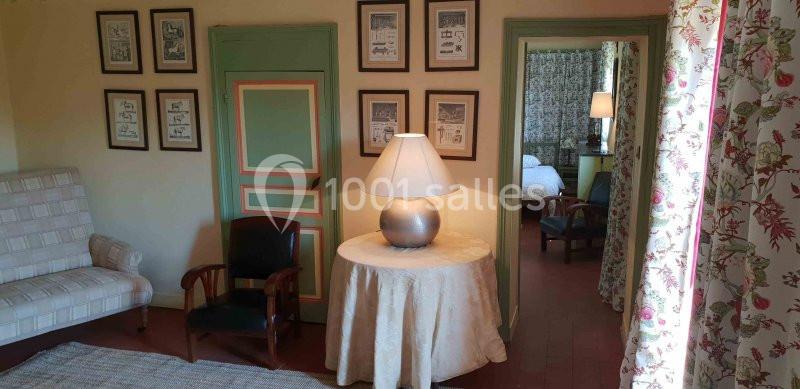 Intérieur d'une pièce avec table ronde, lampe, fauteuils, portes colorées et vue sur une chambre adjacente.