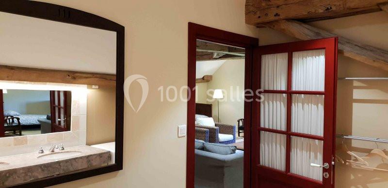 Vue d'une salle de bain avec miroir, lavabo et porte ouverte donnant sur un salon avec canapé et lampe.