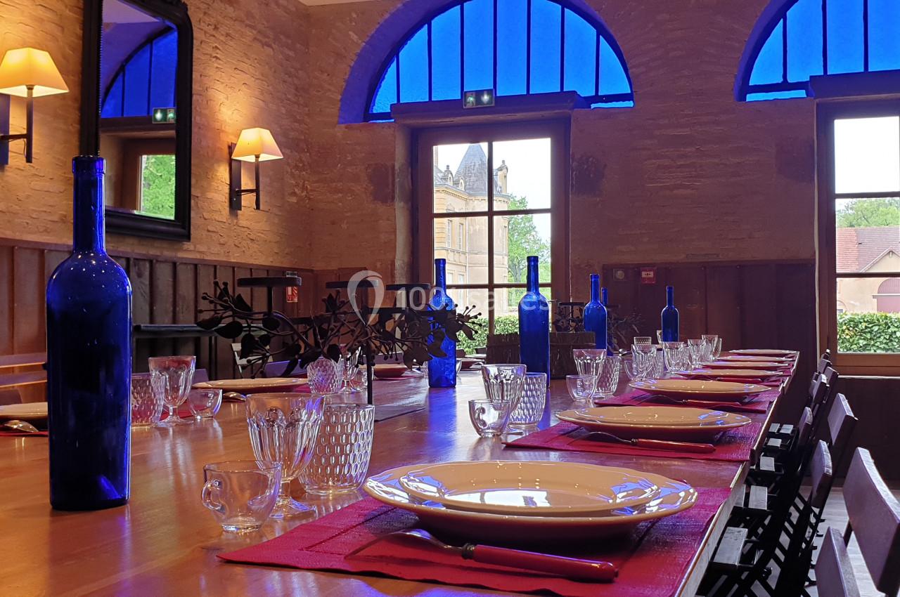 Salle à manger lumineuse avec une grande table en bois dressée, bouteilles bleues et fenêtres donnant sur un jardin.