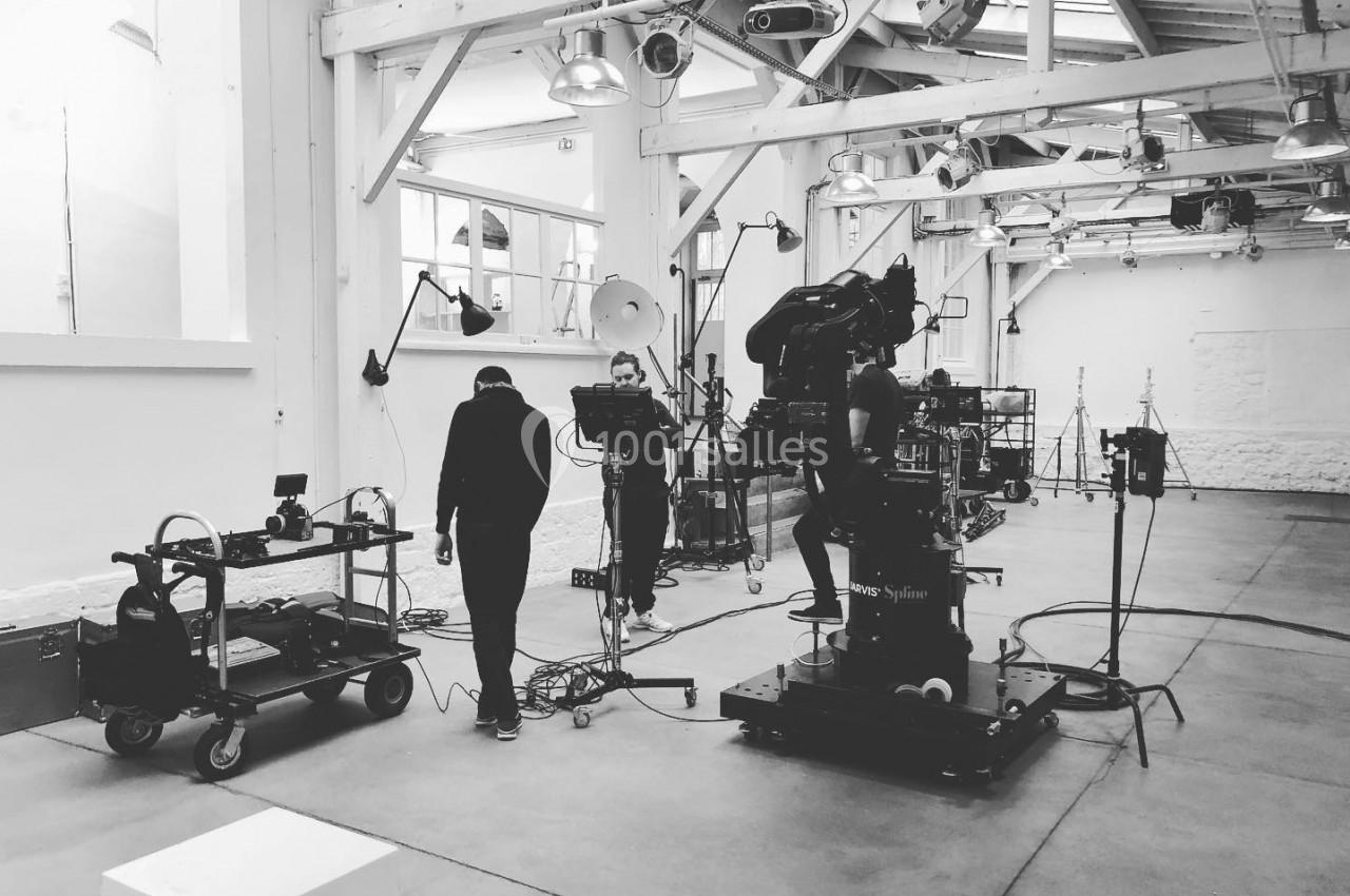 Des techniciens préparent du matériel de tournage dans un studio lumineux avec des structures métalliques apparentes.