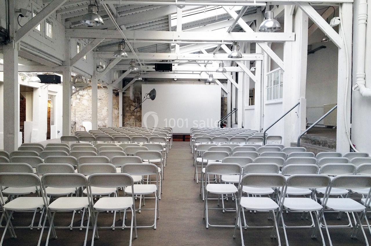 Salle lumineuse avec des rangées de chaises blanches alignées face à un écran de projection dans un espace industriel.