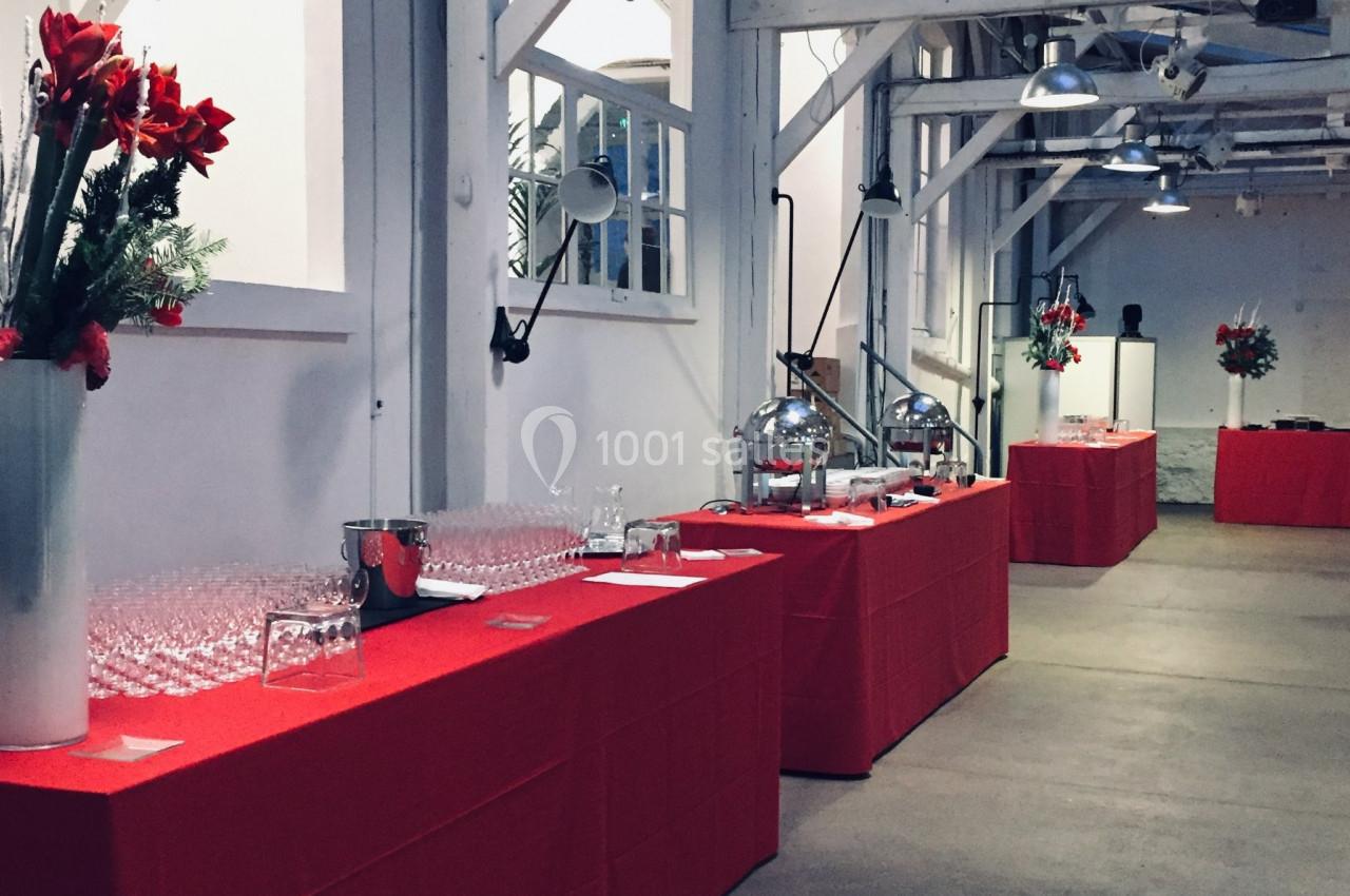 Salle aménagée pour un événement avec tables recouvertes de nappes rouges, verres, fleurs et équipements de service.