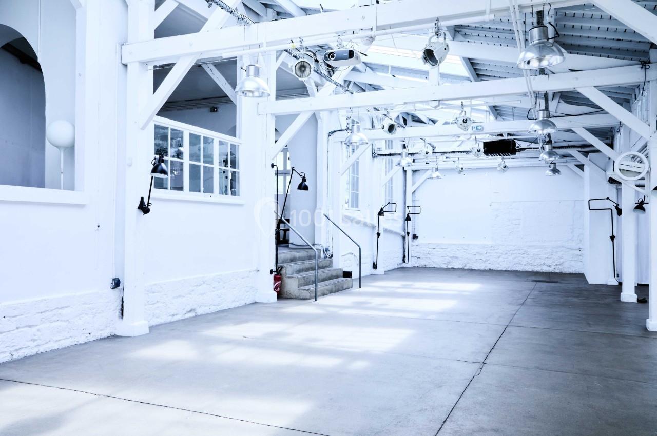 Intérieur d'un grand espace lumineux avec des poutres blanches, des fenêtres et des lampes suspendues.