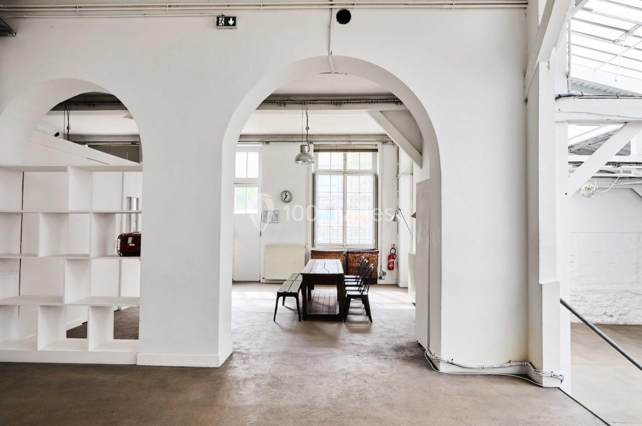 Salle lumineuse avec murs blancs, grande table en bois entourée de chaises, étagères et fenêtres laissant entrer la lumière…