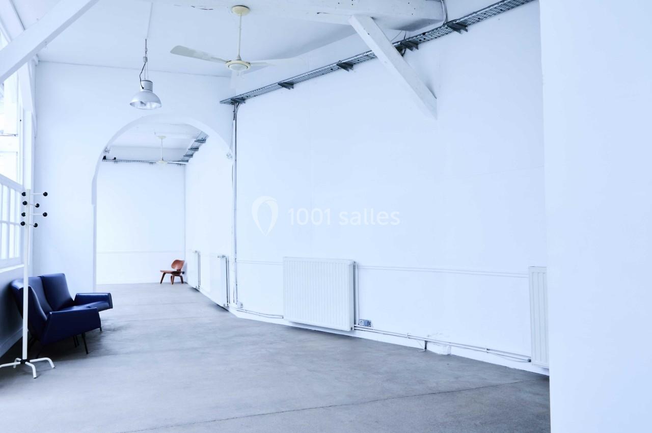 Salle lumineuse avec murs blancs, sol en béton, fauteuils bleus et marron, et poutres apparentes au plafond.