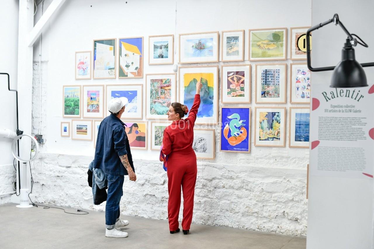Deux personnes regardent et discutent devant un mur d'œuvres d'art encadrées dans une galerie lumineuse.