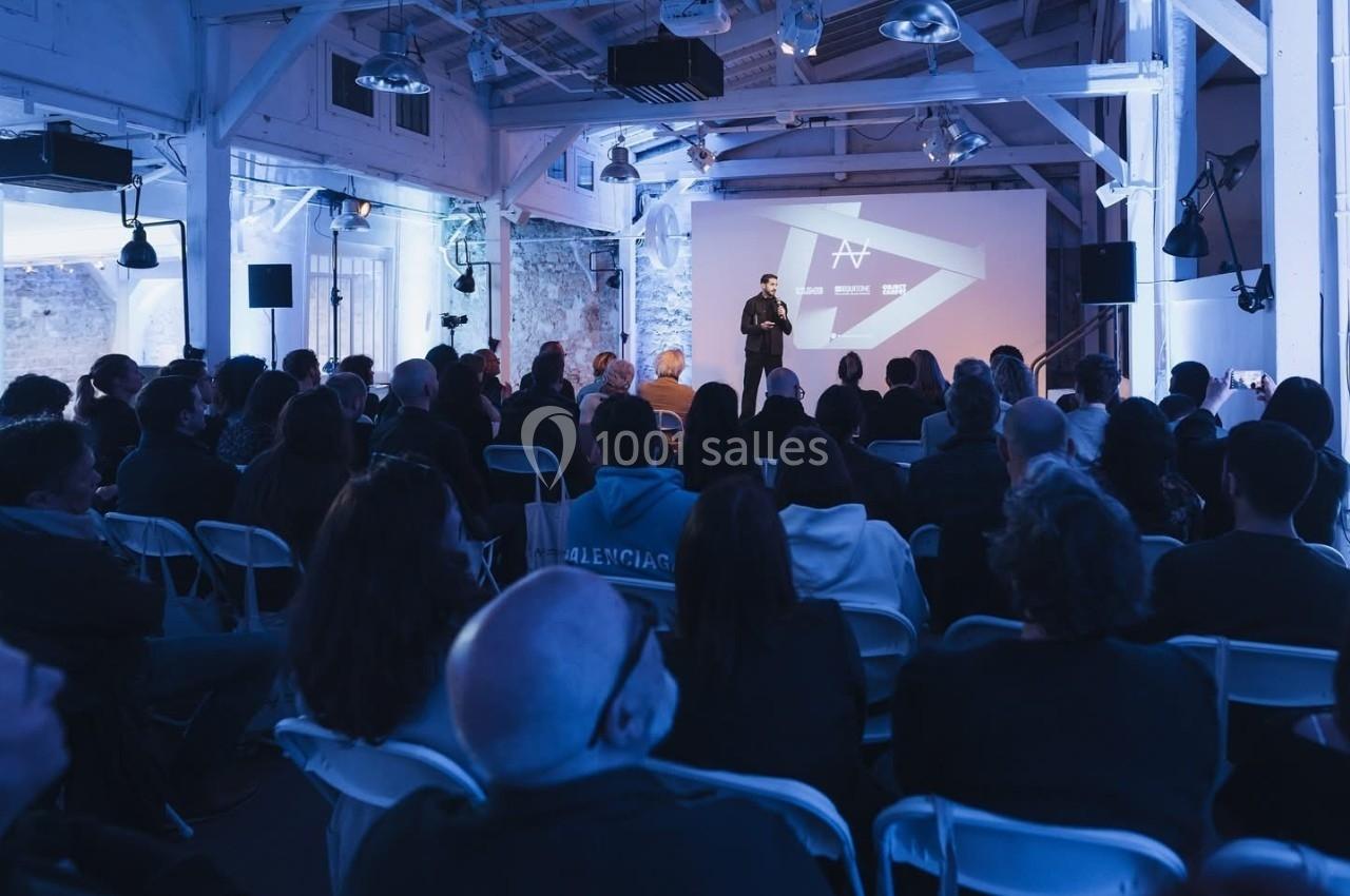 Une personne présente sur scène devant un public assis dans une salle éclairée par des lumières bleues.