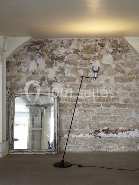 Lampe sur pied inclinée devant un mur en pierre brute, avec un grand miroir appuyé contre le mur.