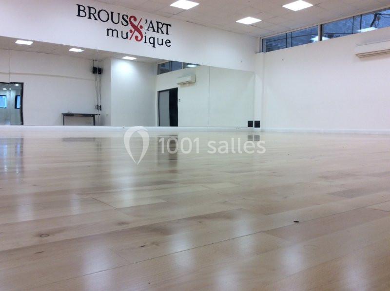 Salle de danse lumineuse avec parquet clair, murs blancs, miroirs et logo ’Brousse Art Musique’ sur un mur.
