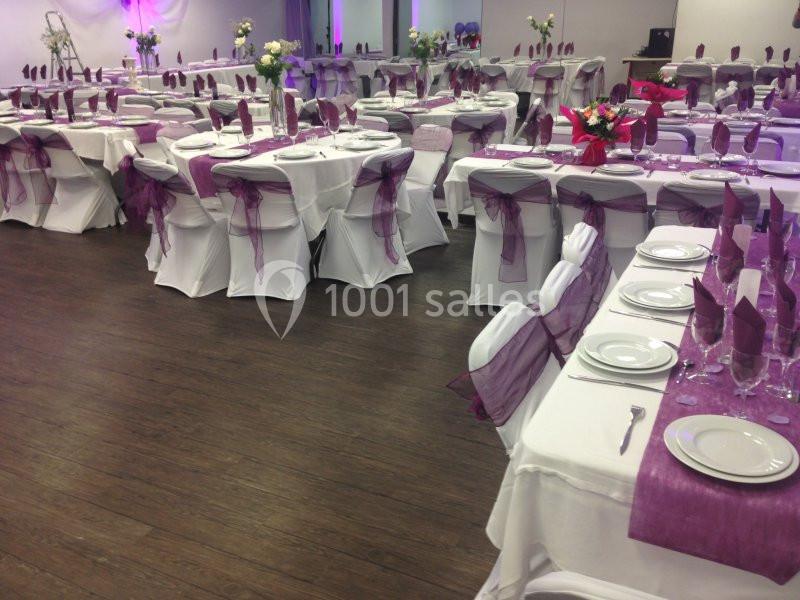 Salle de réception décorée avec des tables dressées, nappes blanches et nœuds violets sur les chaises.