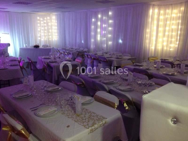 Salle de réception décorée avec des nappes blanches, des chaises ornées de rubans dorés et des guirlandes lumineuses.
