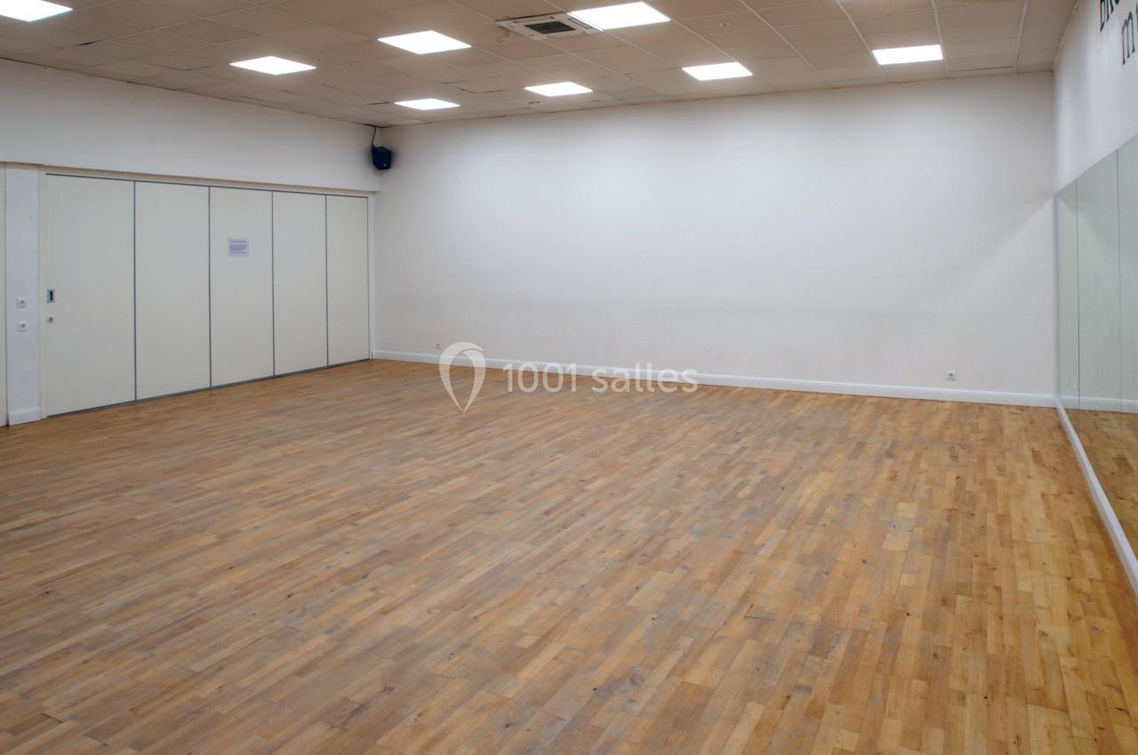 Salle vide avec parquet en bois, murs blancs, plafond éclairé et un grand miroir couvrant un mur latéral.