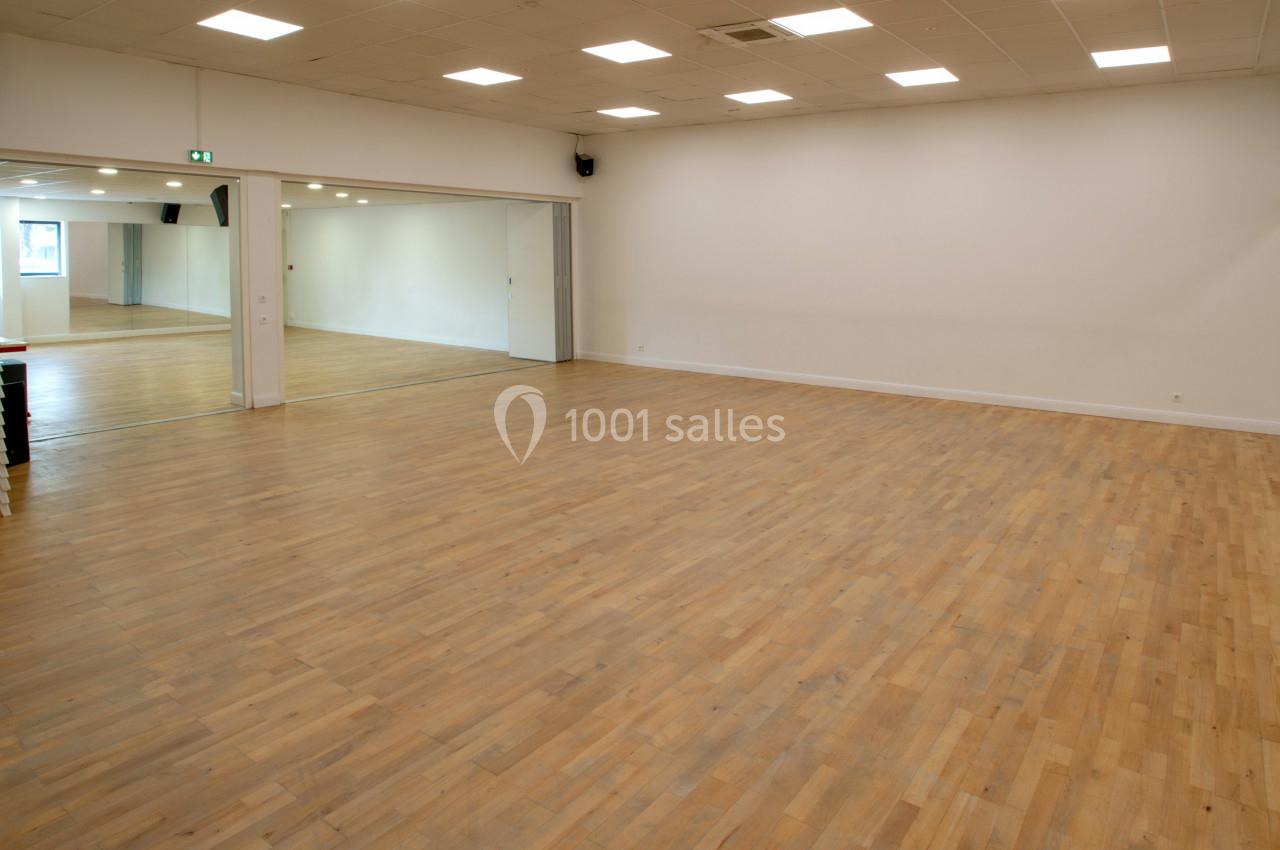 Salle vide avec parquet en bois clair, murs blancs, miroirs sur un mur et chaises empilées dans un coin.