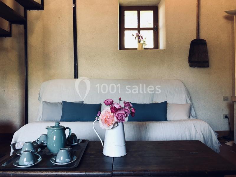 Canapé blanc avec coussins bleus, table en bois avec vase de fleurs et service à thé, intérieur lumineux avec fenêtre.