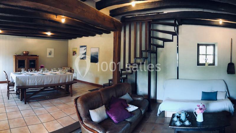 Salle de séjour rustique avec poutres apparentes, table dressée, canapé, escalier en bois et décoration simple.