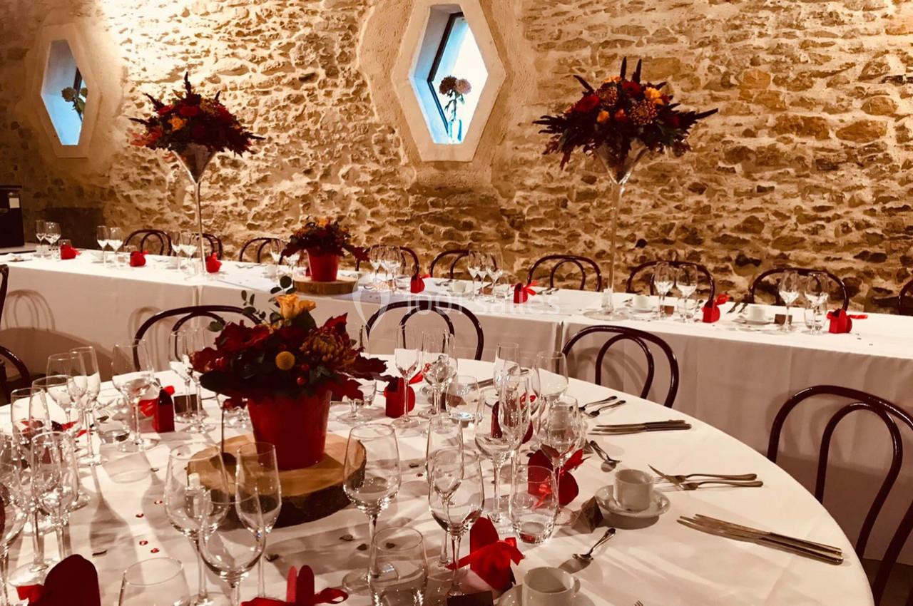 Salle de réception en pierre avec tables dressées, décorations florales rouges et fenêtres hexagonales.