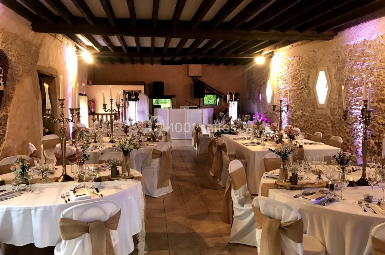Salle de réception décorée pour un mariage, avec tables dressées, nappes blanches et chaises ornées de rubans beige.