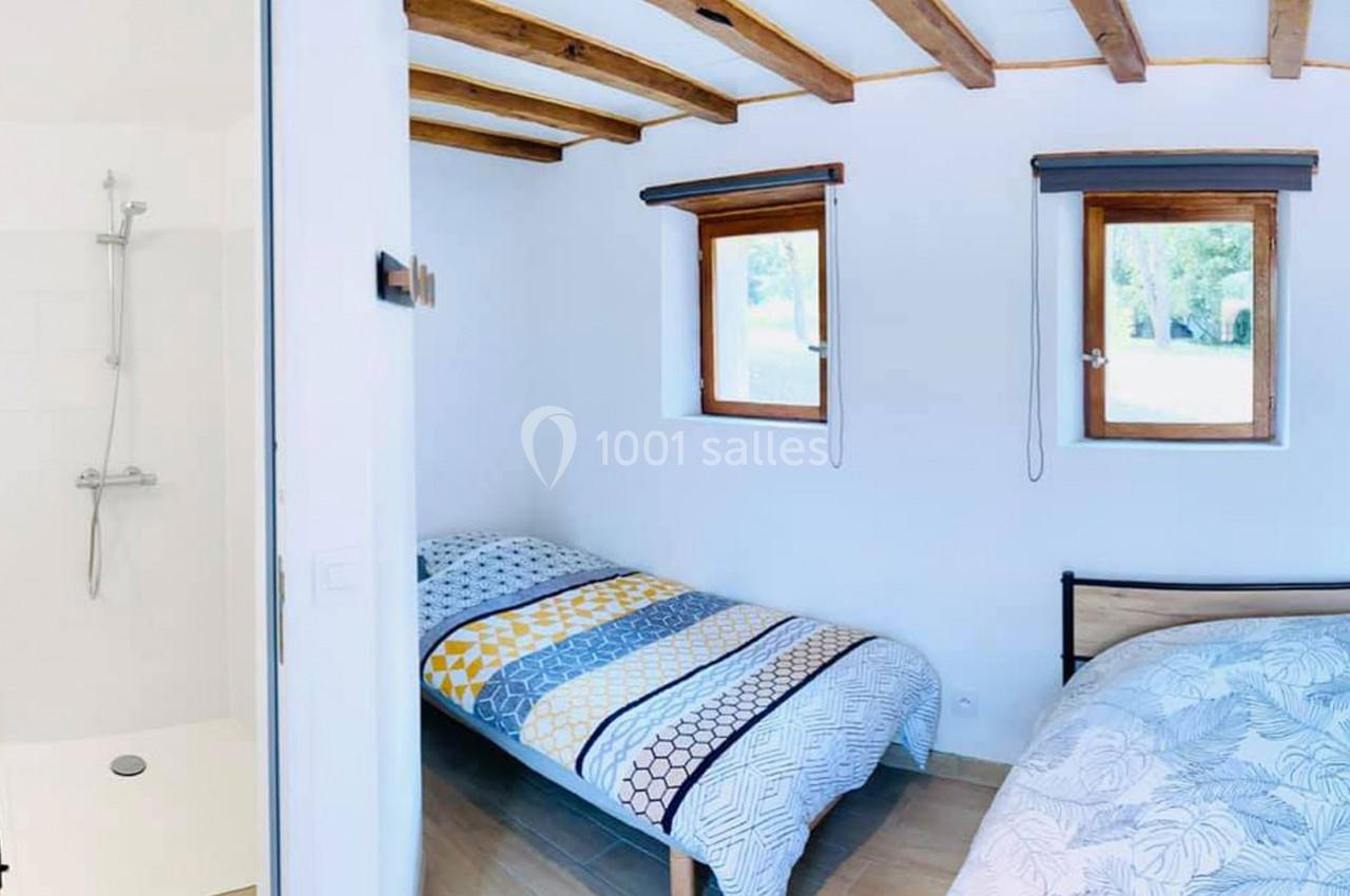 Deux lits simples dans une chambre lumineuse avec plafond en poutres apparentes, vue sur une douche et fenêtres.