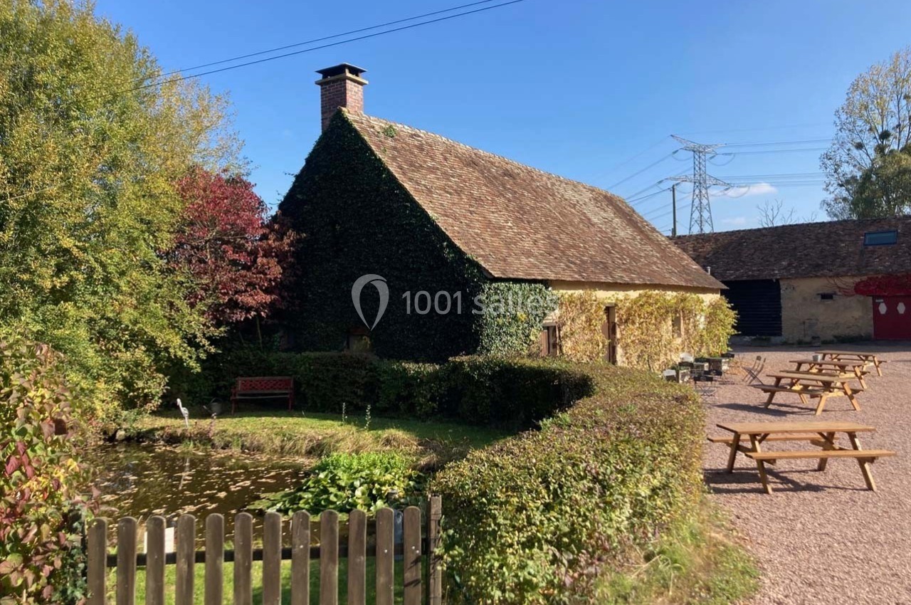 Maison en pierre couverte de lierre, entourée d'un jardin avec un étang, des bancs et des tables en bois.