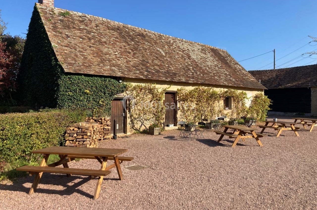 Bâtiment rural en pierre avec toit en tuiles, entouré de végétation, tables de pique-nique sur une cour gravillonnée.