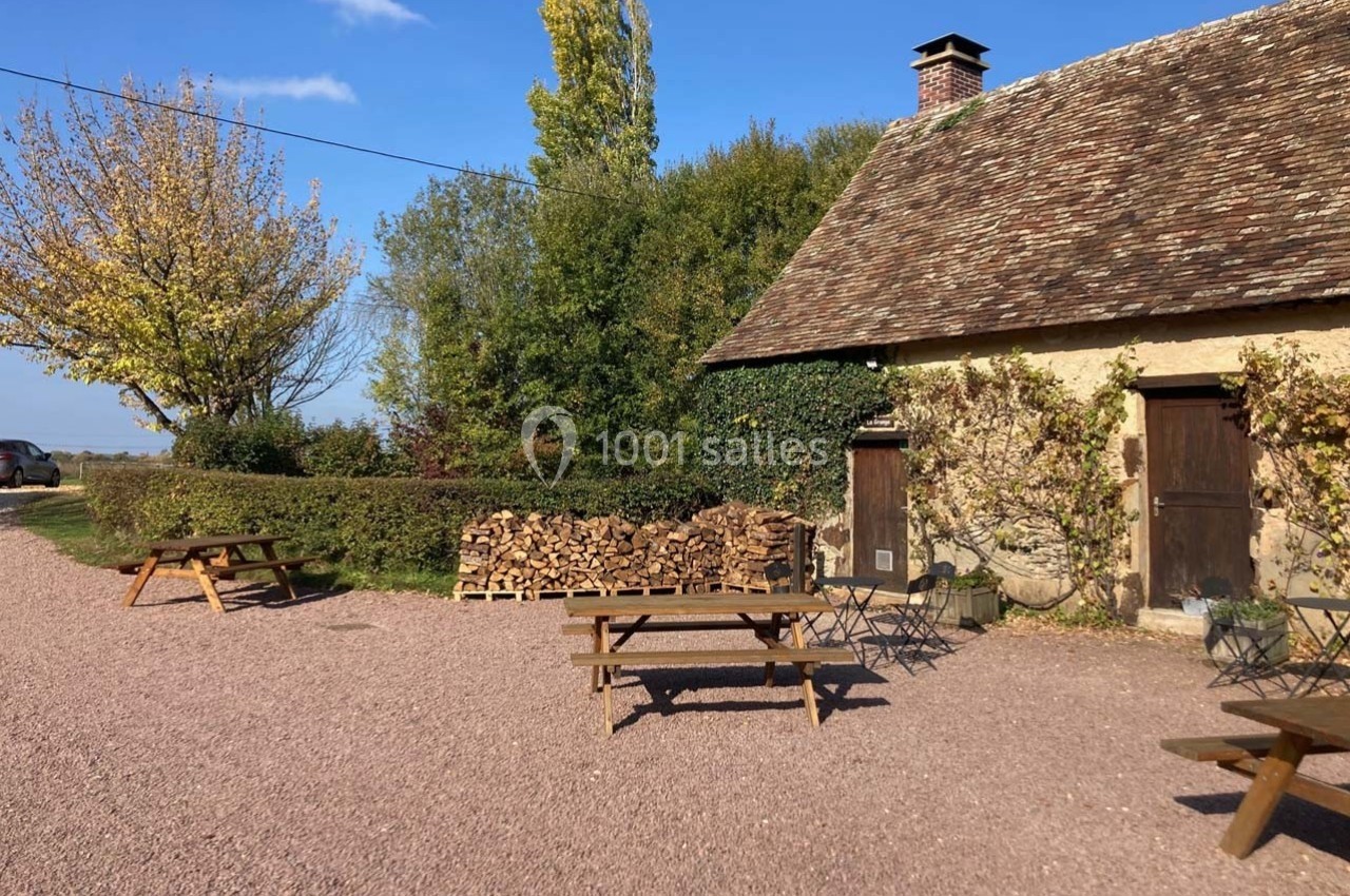 Cour avec tables en bois, chaises et tas de bûches devant un bâtiment en pierre entouré de végétation.