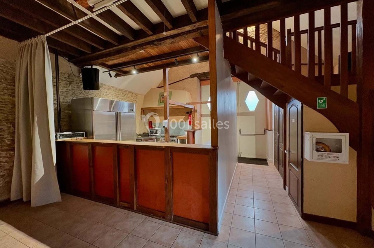 Cuisine ouverte avec comptoir en bois, plafond avec poutres apparentes et escalier en bois menant à un étage.