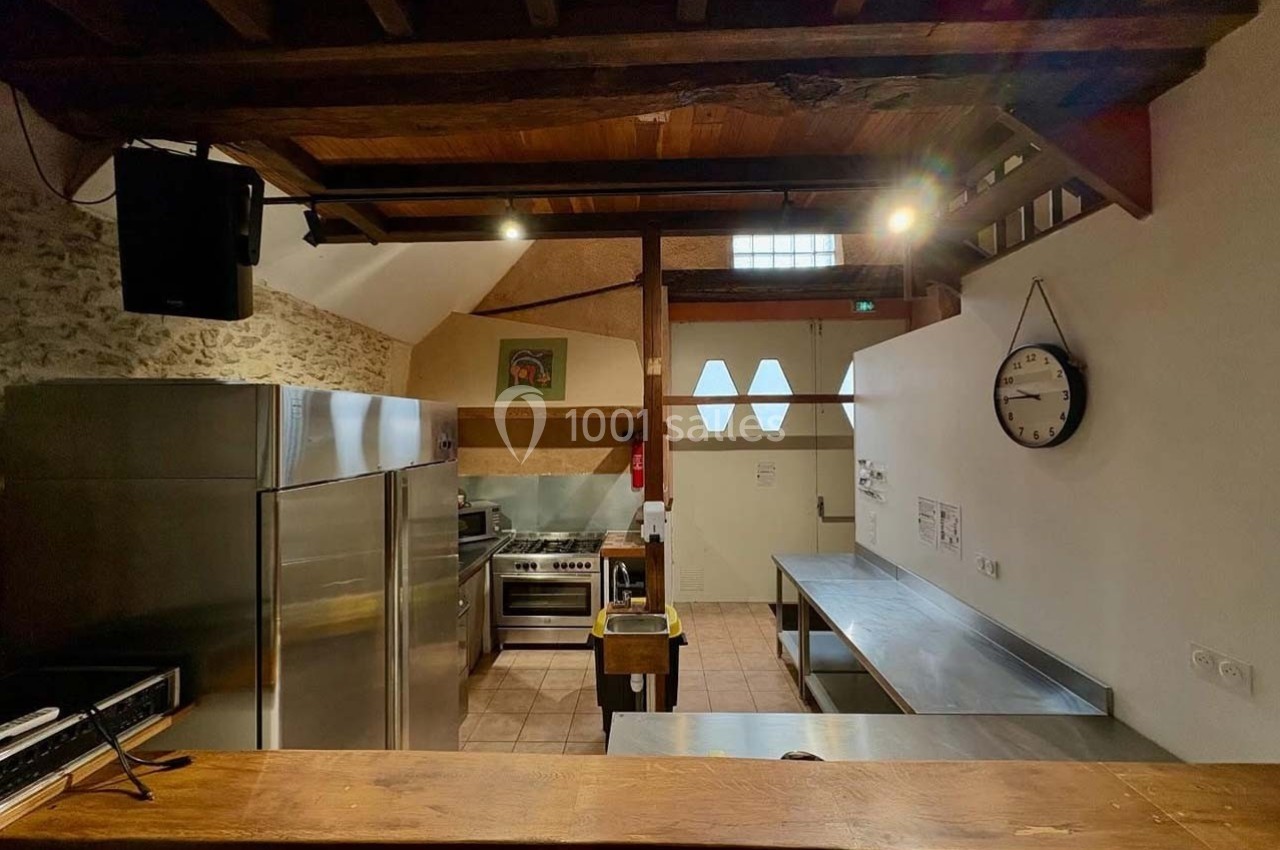 Cuisine spacieuse avec équipements inox, plan de travail, murs en pierre, poutres apparentes et horloge murale.