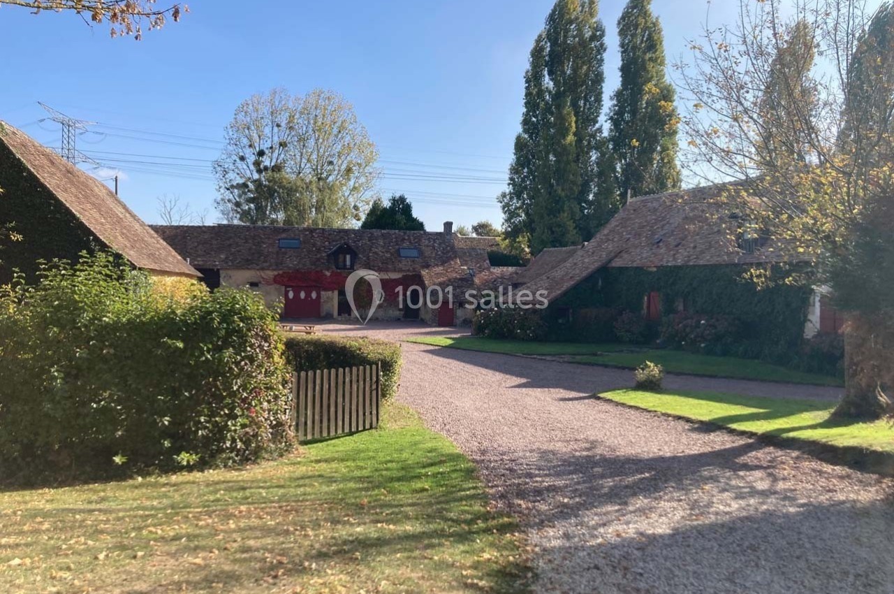Cour d'une ferme traditionnelle avec bâtiments en pierre, toits en tuiles et arbres sous un ciel dégagé.