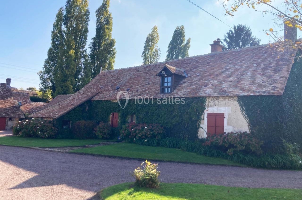Maison ancienne en pierre avec toit en tuiles, façade recouverte de lierre, entourée d'arbres et d'un chemin en gravier.