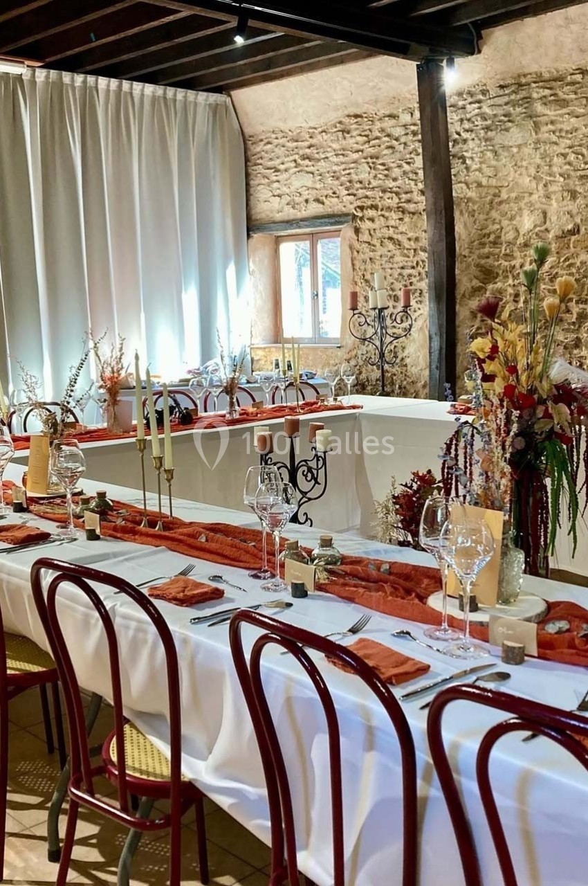 Salle décorée pour un repas, avec une longue table dressée, chaises rouges et ornements floraux aux tons chauds.