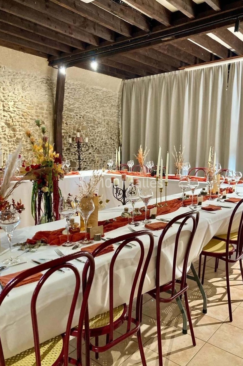 Salle décorée pour un repas, avec tables dressées, chaises rouges, nappes blanches et décorations florales automnales.