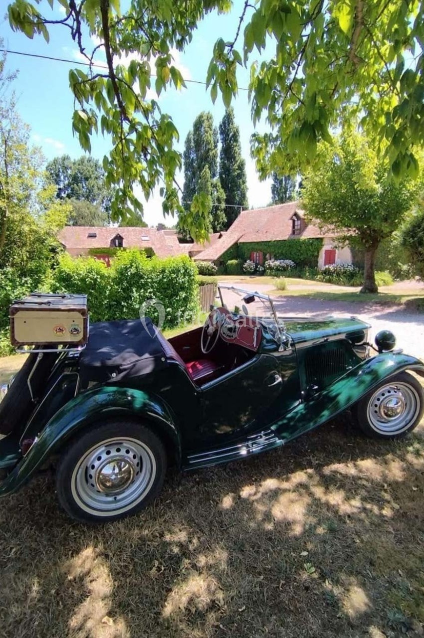 Voiture ancienne verte décapotable garée sous un arbre, avec une maison en briques rouges en arrière-plan.