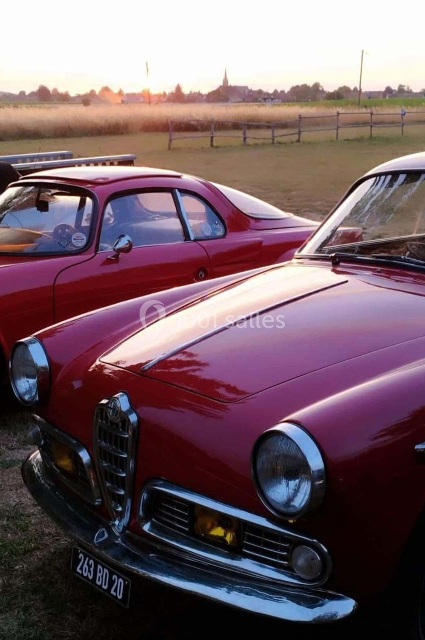 Deux voitures anciennes rouges stationnées dans un champ au coucher du soleil, avec un village en arrière-plan.