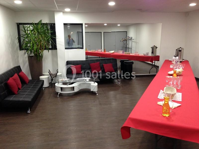 Salle aménagée avec canapés noirs, table basse, plante, miroir mural et table dressée avec nappes rouges et boissons.