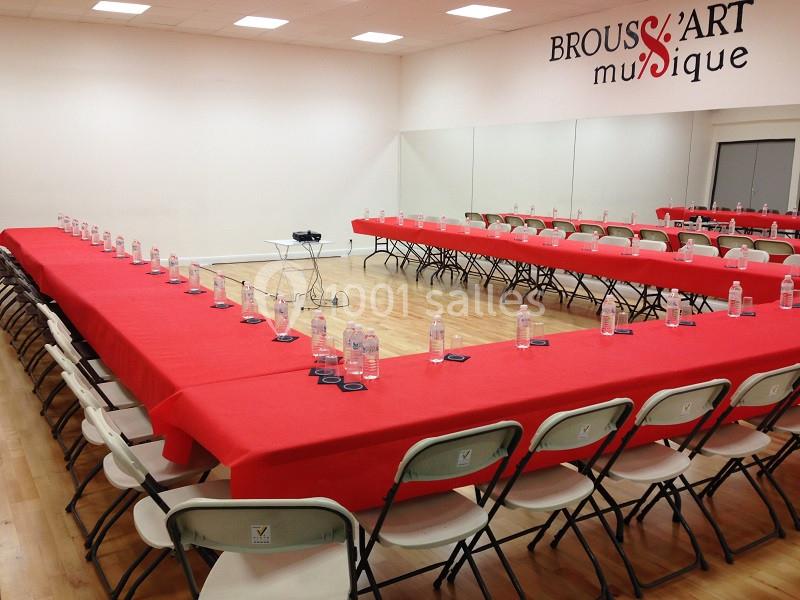 Salle de réunion avec tables en U couvertes de nappes rouges, chaises pliantes et bouteilles d'eau disposées.