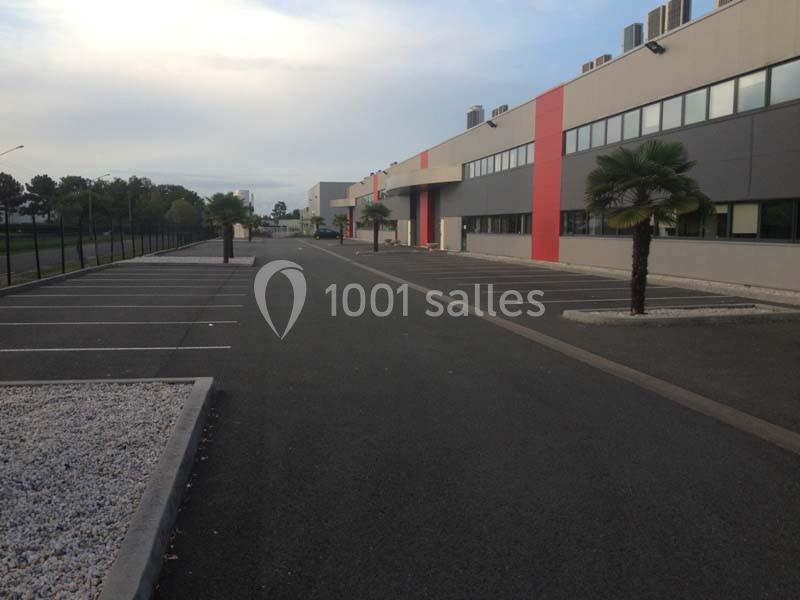 Parking vide devant un bâtiment industriel moderne avec des palmiers et une clôture longeant la route.